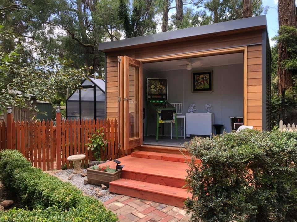 Designer Hideaways Hideout studio with cedar cladding and bifold doors, set in a lush backyard garden with red timber steps, brick path, and decorative landscaping.