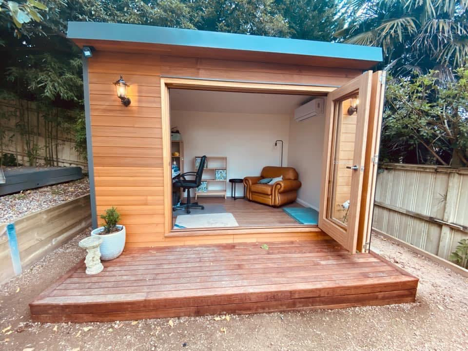 Designer Hideaways Hideout studio set up as a home office with cedar cladding, open bifold doors, timber deck, and modern interior featuring a desk and leather lounge.
