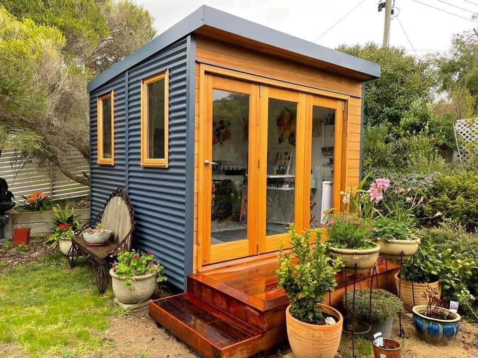 Compact backyard garden studio with cedar cladding and dark Colorbond walls, featuring bifold timber doors and surrounded by potted plants — ideal for a home office or creative retreat.