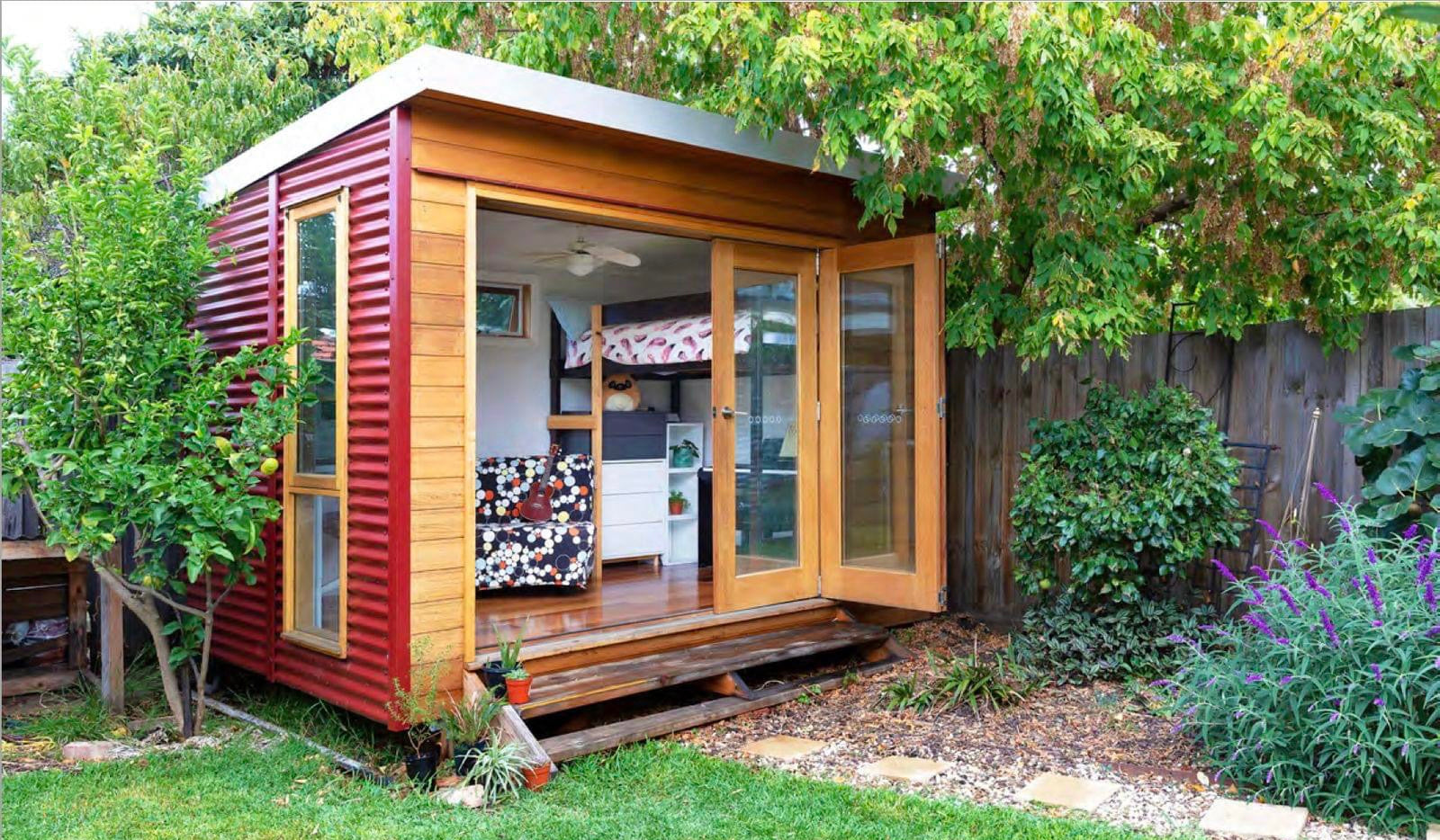 Designer Hideaways Hideout studio set up as a cozy bunk room with cedar cladding, Manor Red Colorbond walls, open timber bifold doors, and elevated timber steps nestled in a leafy backyard.